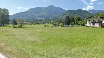 Weitläufige grüne Wiese mit Bergblick und ländlicher Umgebung unter blauem Himmel.