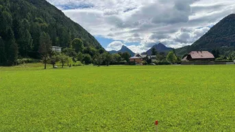 Weitläufige grüne Wiesenlandschaft mit beeindruckendem Bergblick und Häusern in der Ferne.