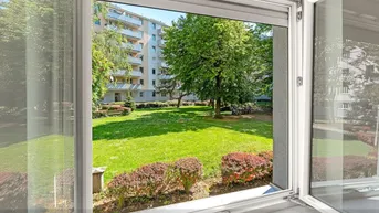 Blick aus einem geöffneten Fenster auf einen grünen Innenhof mit Bäumen und Rasenflächen.