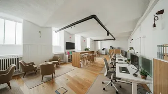 Geräumiges, modernes Büro mit Holzboden, Arbeitsplätzen, Lounge-Bereich und sichtbaren schwarzen Rohren an der Decke.