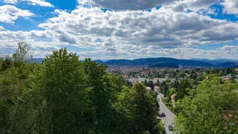 Panoramablick über eine Stadtlandschaft mit grünen Hügeln und Bergen im Hintergrund unter blauem Himmel.