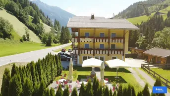 Panoramablick auf ein gepflegtes Gebäude im alpinen Stil mit mehreren Balkonen und einer Außenterrasse, umgeben von grünen Bergen und Wiesen.