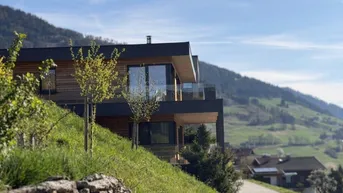 Modernes Haus in Hanglage mit Holzfassade, großen Fenstern und Panoramablick auf die Berge.