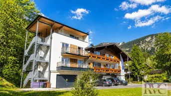 Modernes Gebäude mit traditionellen Holzelementen und Balkonen vor alpiner Bergkulisse im Sommer.