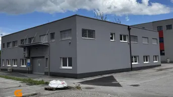 Modernes graues Gebäude mit vielen Fenstern und einem gepflasterten Vorplatz unter bewölktem Himmel.