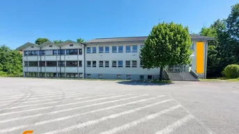 Großes Bürogebäude mit weitläufigem Parkplatz und grüner Umgebung unter blauem Himmel.
