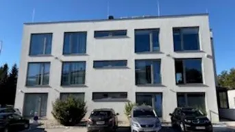 Modernes, weißes Gebäude mit großen Fenstern und Parkplätzen davor unter blauem Himmel.