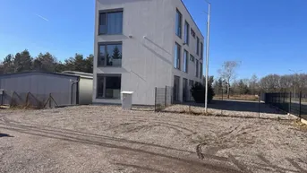 Modernes, weißes Gebäude mit großen Fenstern und einem Schotterparkplatz unter blauem Himmel.