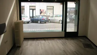 Leerer Raum mit großen Fenstern zur Straße, ausgestattet mit einem Heizkörper und Holzplankenboden.