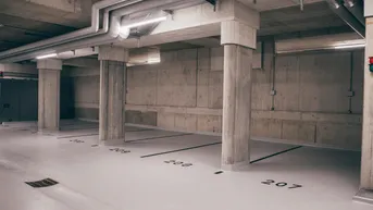 Moderne Tiefgarage mit nummerierten Stellplätzen, Betonwänden und sichtbaren Installationen an der Decke.