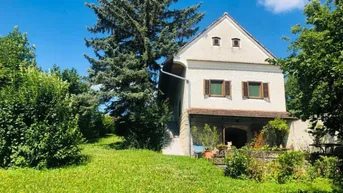 Traditionelles Landhaus mit großem Garten und altem Baumbestand unter blauem Himmel.