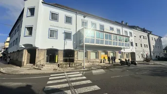 Mehrstöckiges Gebäude mit hellblauer Fassade, großen Fenstern und einem überdachten Balkon an einer belebten Straßenecke.