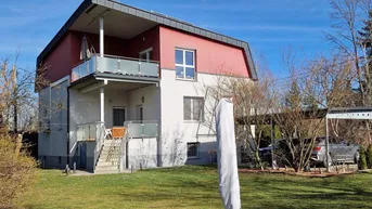 Modernes Wohnhaus mit weißer und roter Fassade, Balkon mit Glasgeländer und einem gepflegten Garten.