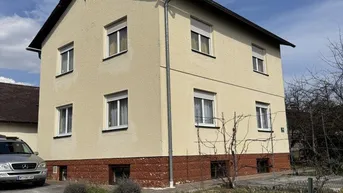 Einfamilienhaus mit heller Fassade, Satteldach und Vorgartenbereich unter blauem Himmel.