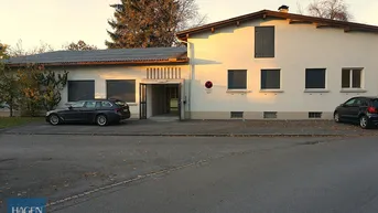 Gebäudeansicht mit Parkplätzen und Bäumen im Herbstlicht, das die Fassade beleuchtet.