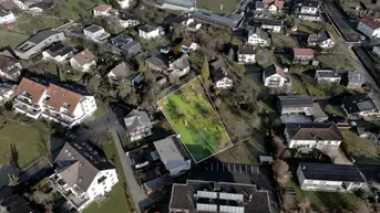 Luftaufnahme eines Baugrundstücks mit umliegenden Wohnhäusern und einem Sportplatz im Hintergrund.