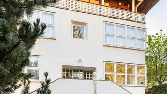 Weißes Gebäude mit moderner Architektur, großen Fenstern, Balkon und gepflegtem Garten.