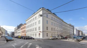 Weißes Eckgebäude mit klassischer Architektur an einer städtischen Straße unter blauem Himmel.