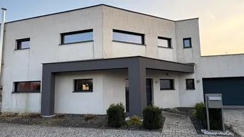 Modernes Einfamilienhaus mit heller Fassade, Flachdach und überdachtem Eingangsbereich, umgeben von gepflasterter Auffahrt und Kiesfläche.
