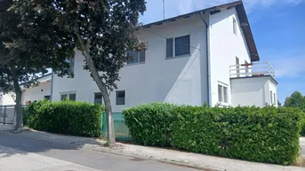 Weißes Wohnhaus mit grüner Hecke und Bäumen an einer Straße unter blauem Himmel.