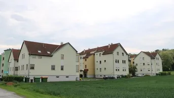 Reihenhausanlage mit hellen Fassaden und roten Ziegeldächern, umgeben von grünen Feldern unter bewölktem Himmel.