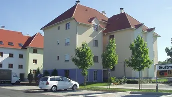 Mehrfamilienhaus mit heller Fassade, rotem Ziegeldach und gepflegter Außenanlage mit Parkplätzen.