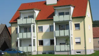 Mehrfamilienhaus mit gelber Fassade, rotem Ziegeldach und mehreren Balkonen, umgeben von Grünflächen.
