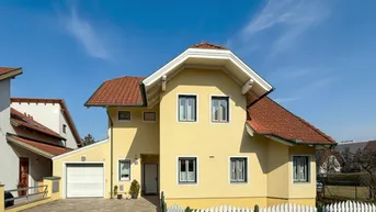Gelbes Einfamilienhaus mit rotem Ziegeldach, weißem Zaun und integrierter Garage unter blauem Himmel.