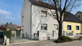 Vorderansicht eines hellgrauen Einfamilienhauses mit gepflasterter Einfahrt und einem Baum davor.