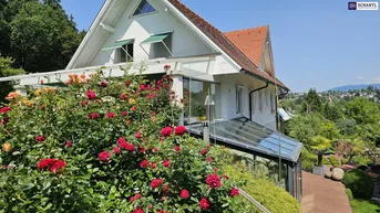 Hausfassade mit blühenden Rosen und einem Wintergarten, umgeben von üppigem Grün und Fernblick.