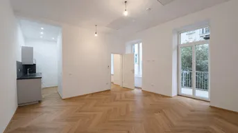 Heller Wohnbereich mit Fischgrätparkett, hohen Decken und integrierter Küchenzeile sowie Zugang zu einem französischen Balkon.