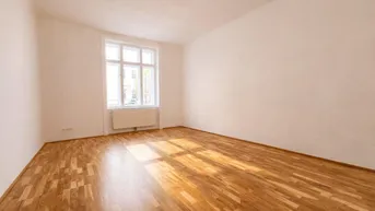 Helles, leeres Zimmer mit Parkettboden und großem Fenster, das viel Tageslicht hereinlässt.