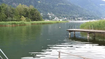 Malerischer Blick auf den See mit bewaldeten Bergen im Hintergrund und einem kleinen Steg.
