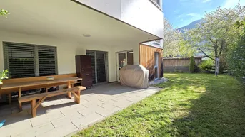Geräumige Terrasse mit Holztisch und Gartenbereich, umgeben von grüner Hecke und Bergblick.