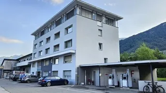 Modernes Bürogebäude mit mehreren Etagen, Parkplätzen und einer Tankstelle im Vordergrund, umgeben von Bergen.