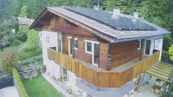 Traditionelles Haus mit Holzfassade, Balkon und Solaranlage auf dem Dach, umgeben von viel Grün.