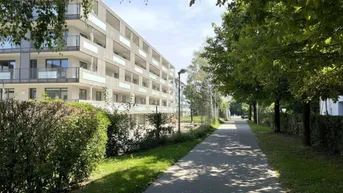 Modernes Mehrfamilienhaus mit zahlreichen Balkonen, umgeben von üppigem Grün und einem sonnigen Spazierweg.