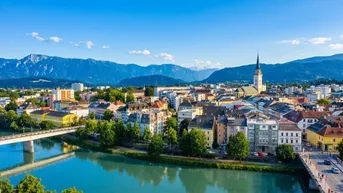 Panoramablick auf eine malerische Stadt mit Fluss, Brücken und majestätischen Bergen im Hintergrund unter blauem Himmel.