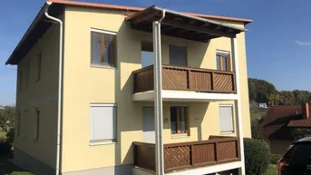 Gelbes Mehrfamilienhaus mit zwei übereinanderliegenden Holzbalconen und grüner Umgebung.