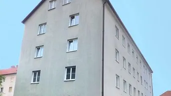 Mehrfamilienhaus mit heller Fassade, Satteldach und Fensterläden, umgeben von grüner Hecke.