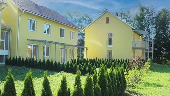 Zwei moderne gelbe Wohnhäuser mit gepflegtem Garten, Hecken und Balkonen unter blauem Himmel.