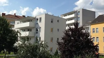 Modernes Mehrfamilienhaus mit hellgrauer Fassade, mehreren Balkonen und umgebender Grünfläche unter blauem Himmel.