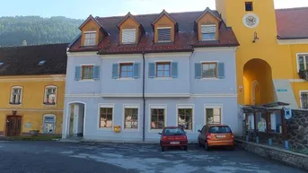 Gebäude mit hellblauer Fassade, Dachgauben und Blick auf die Berge, gelegen an einem Dorfplatz.