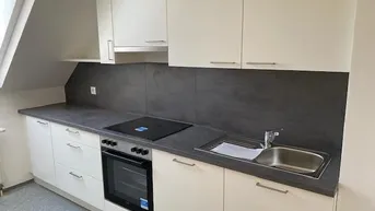 Moderne Einbauküche mit weißen Schränken, dunkler Arbeitsplatte und integrierten Geräten unter Dachschräge.