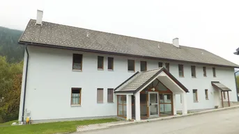 Weißes Mehrfamilienhaus mit Satteldach und überdachtem Eingangsbereich in ländlicher Umgebung.