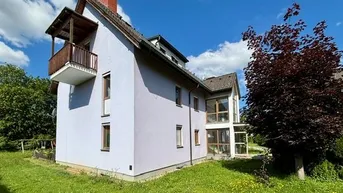 Freistehendes Einfamilienhaus mit hellvioletter Fassade, Holzbalkon und großem Gartenbereich unter blauem Himmel.