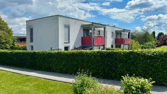 Modernes Gebäude mit weißer Fassade und roten Balkonen, umgeben von einer grünen Hecke unter blauem Himmel.