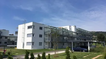 Modernes Mehrfamilienhaus mit Balkonen, Carports und Grünflächen unter blauem Himmel.