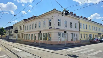 Eckgebäude an einer belebten Kreuzung mit Straßenbahnschienen und hellem Putz unter blauem Himmel.
