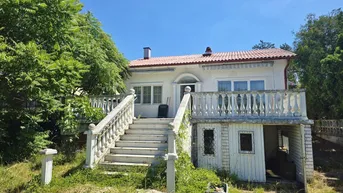 Freistehendes Haus mit markanter Außentreppe, Balkon und mediterranem Ziegeldach unter blauem Himmel.